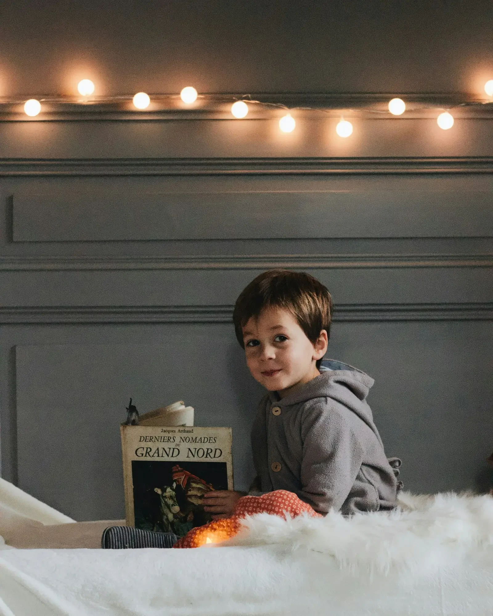 A young boy sitting up in bed with a storybook, under a string of glowing fairy lights.