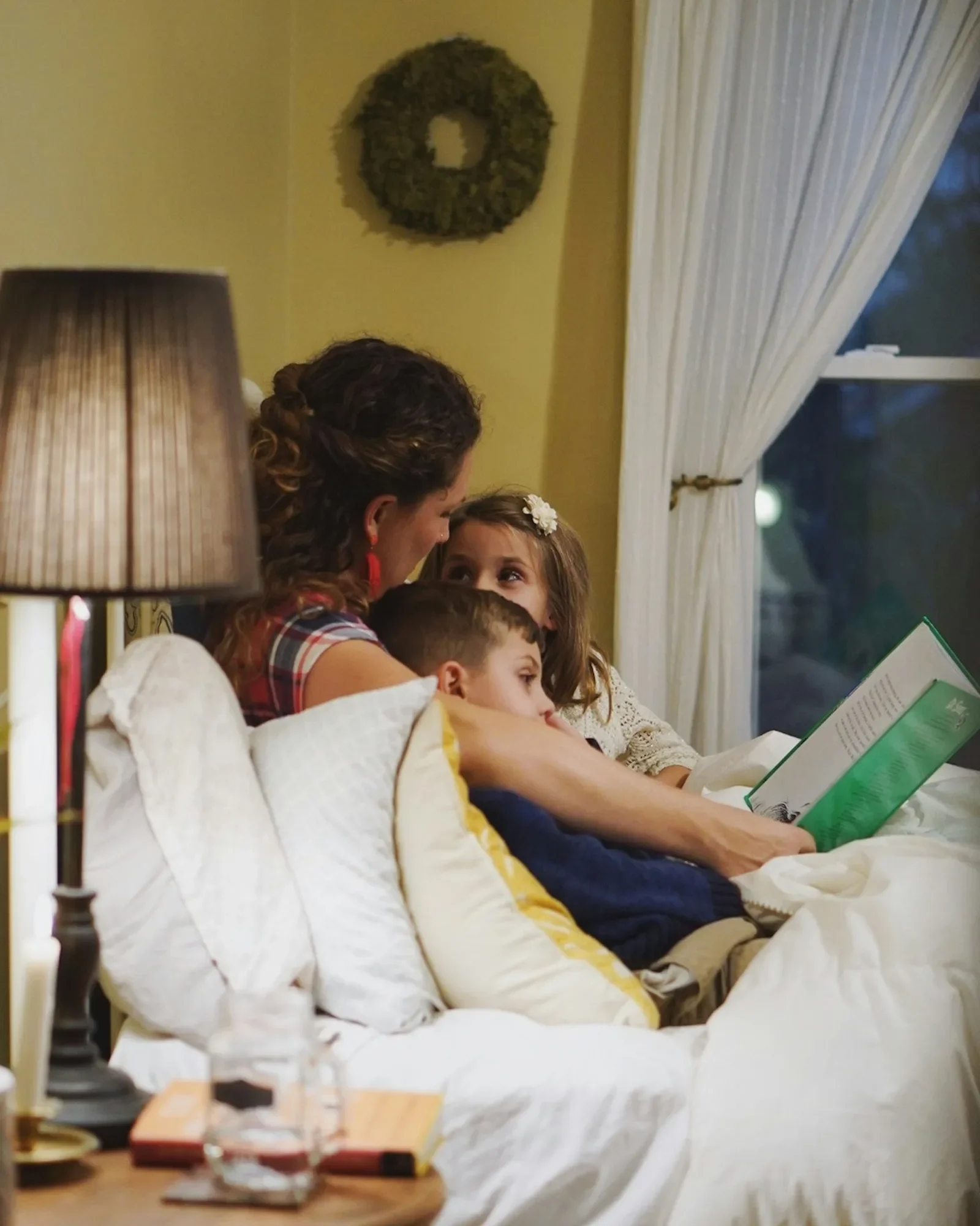 A mother reading a bedtime story to two children tucked up in bed beside a warm lamp.